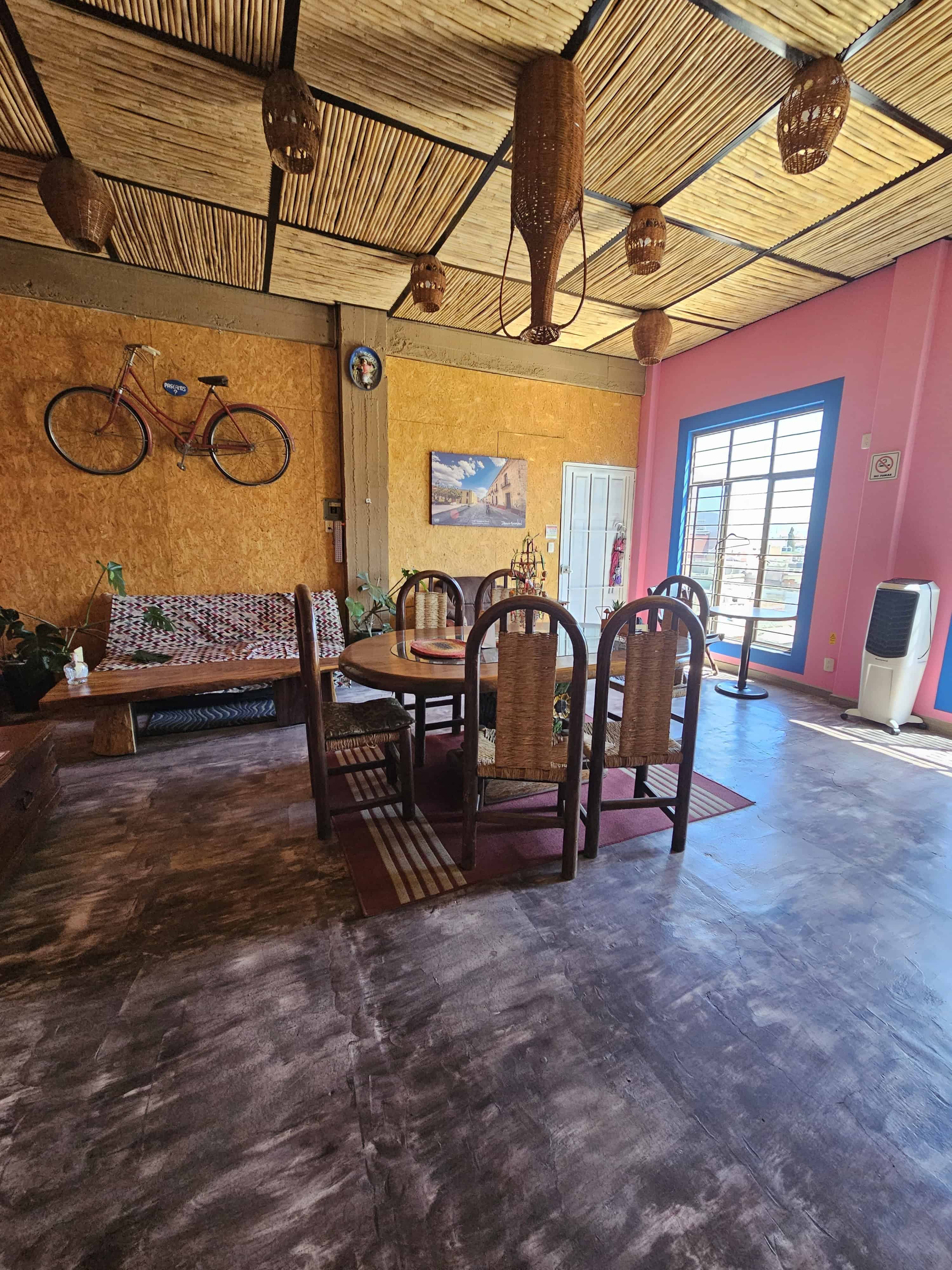 Dining Room view of Rooms by Diaza in Oaxaca, Mexico. Yellow and pinkish orange walls, grass cloth squares on the ceiling with pottery hanging down, bike hanging on the wall, large window outlined in blue and dining table