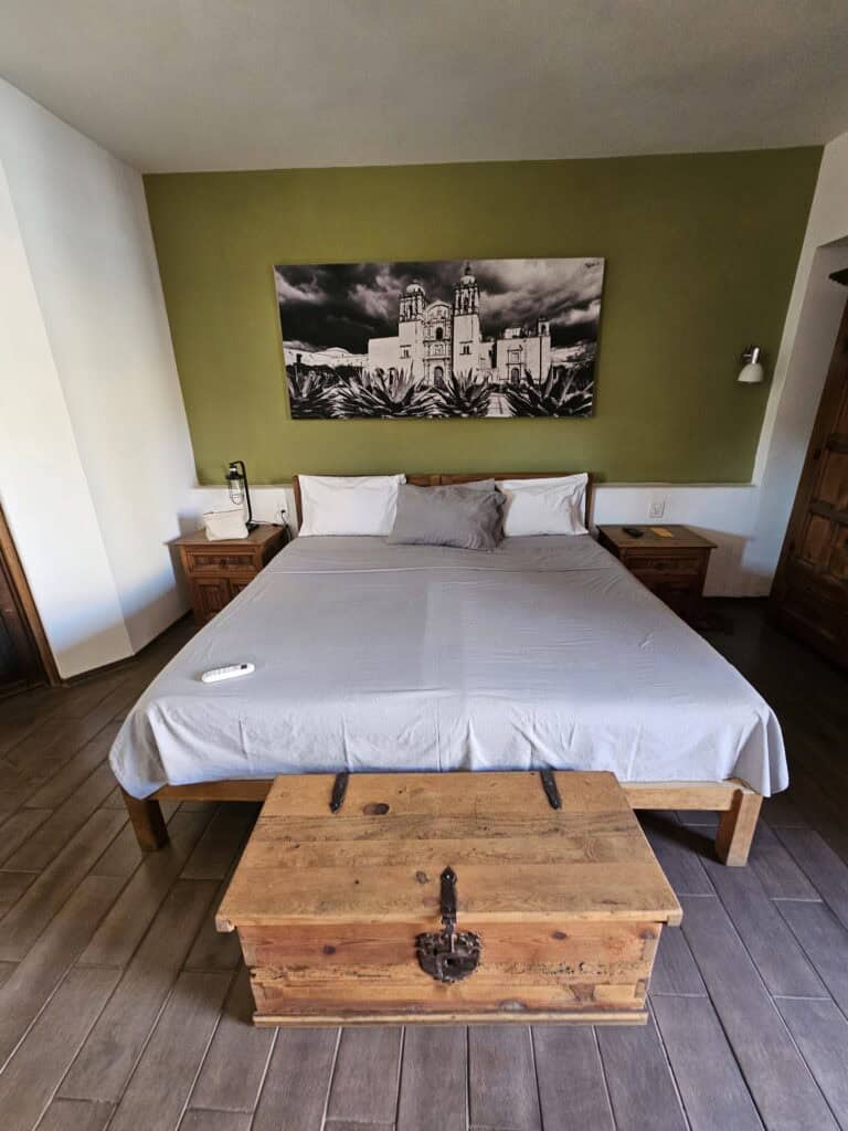 Interior room view of a hotel in Oaxaca Mexico. A bed with white bedding, wooden chest at the foot, green wall behind the bed and a black and white photo above the bed.