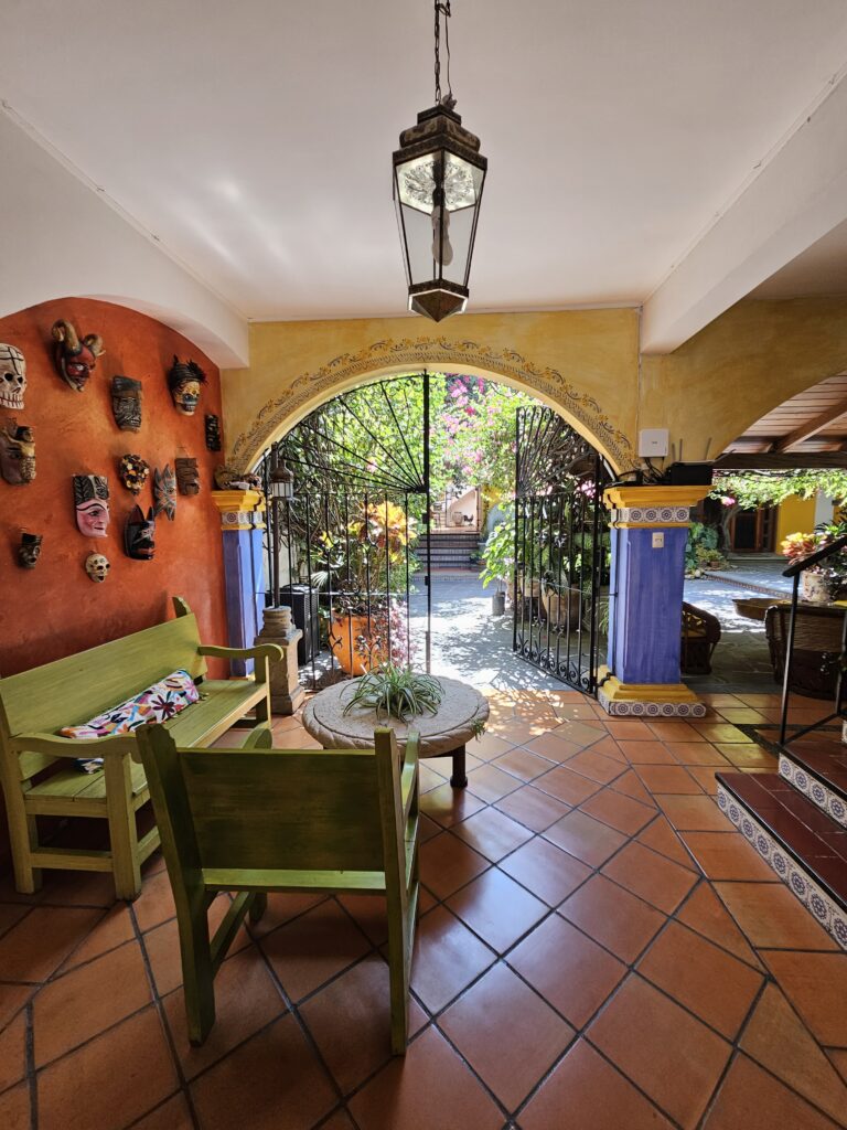 Entrance view of B&B, La Casa Mis Recuerdos, in Oaxaca Mexico, orange tile floors, bright green sofa, orange wall decorated with masks with a view through the wrought iron gate into the courtyard.