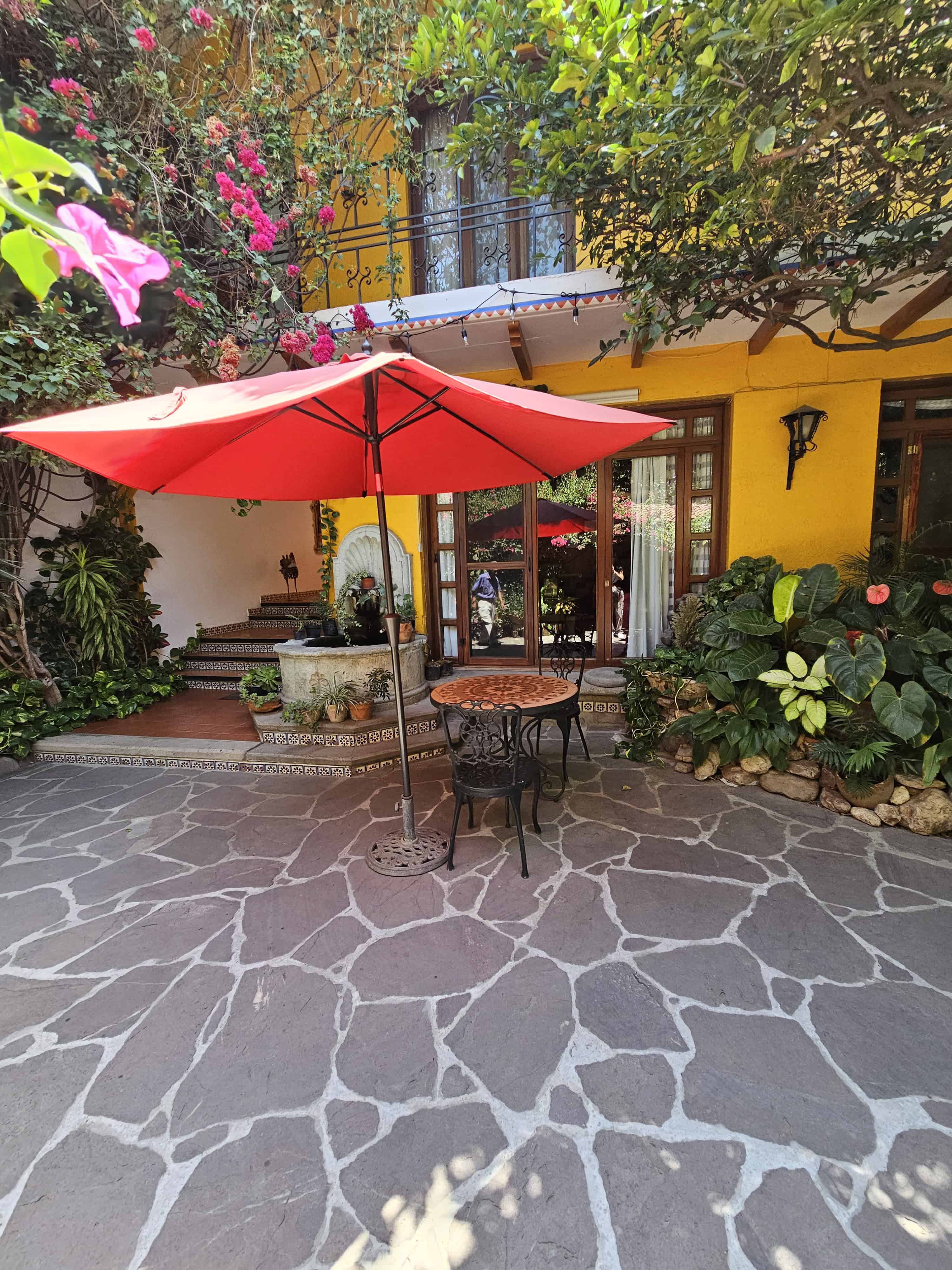Courtyard view at La Casa de Mis Recuerdos B&B in Oaxaca Mexico a metal table and chairs with a bright red umbrella against the yellow walls surrounded by greenery and flowers