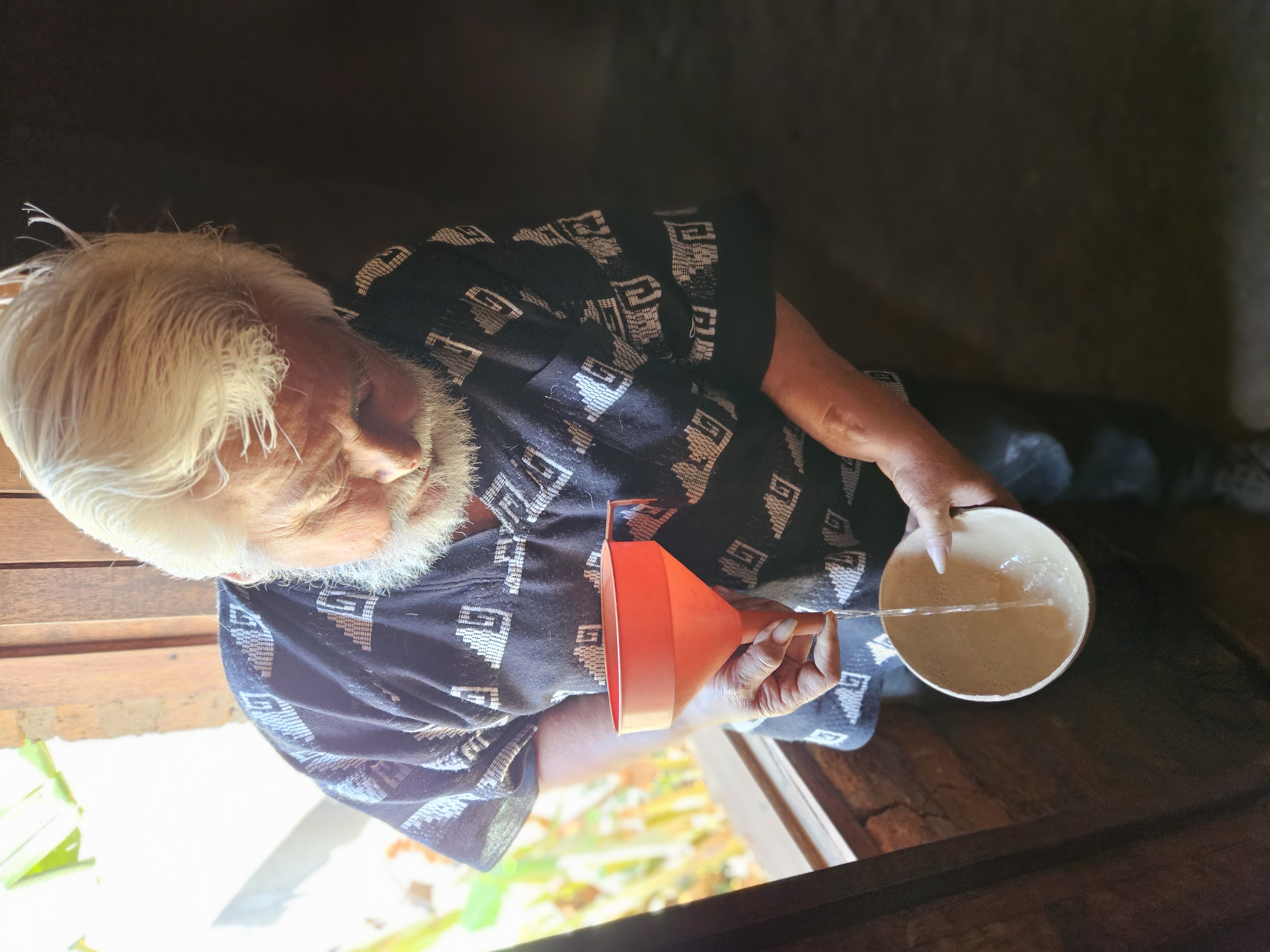 Owner of the palenque, where mezcal is made, standing in front of an open window pouring Mexcal through a red funnel into a traditional gourd dish for tasting in Oaxaca Mexico