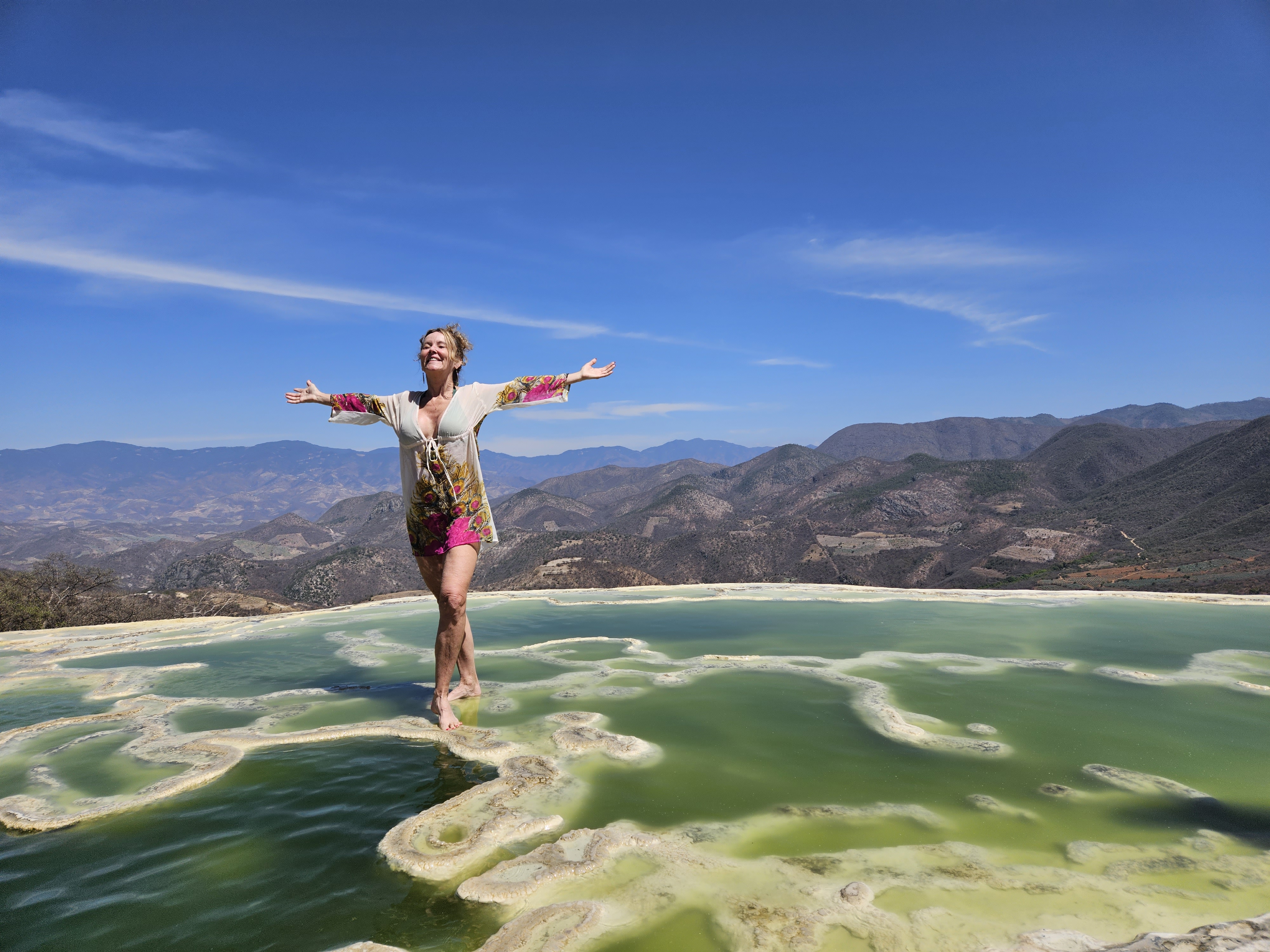 Photo of me with arms outstretched standing in the vibrant green pools of water at Hierve el Agua with a backdrop of the brilliant blue sky in Oaxaca Mexico