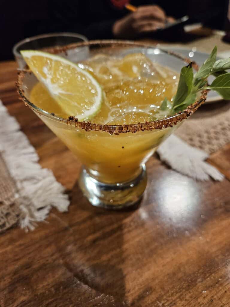 A yellow cocktail of Mezcal with a orange slice and basil