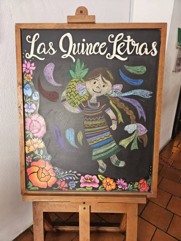 Chalkboard signage for Las Quince Letras restaurant in Oaxaca Mexico with colorful flowers around a young girl holding a pineapple.