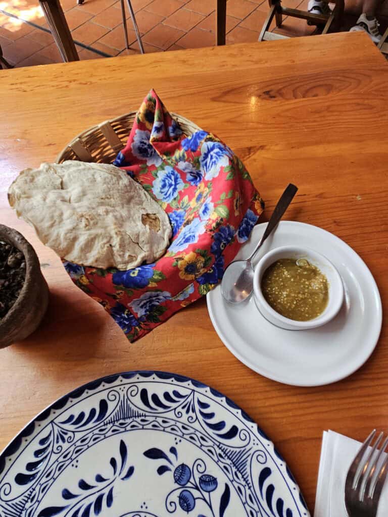 Bread in a basket on a colorful red and blue towel with a side of sauce and colorful blue and white plate.