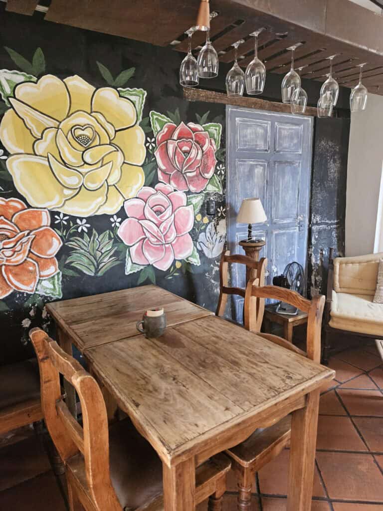 Interior view of Jalatlaco Cafe in Oaxaca, wood table and chairs with a backdrop of large flowers in chalk in colors of yellow, orange, pink and red