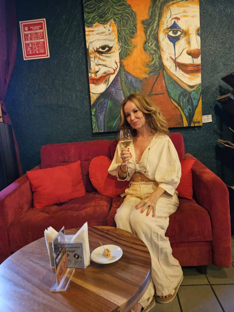 Me sitting on a red sofa with a glass of white wine in my hand, the artwork behind me is the joker. This is at Vino & Vinyl in Oaxaca Mexico