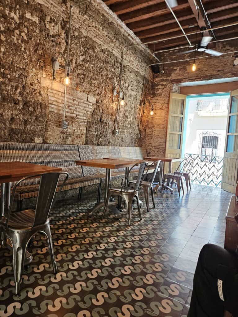 Interior view of Sabina Sabe with tile floors, a rough rock wall, bench seating along the wall with chairs and tables looking out to the open door to the street in Oaxaca Mexico