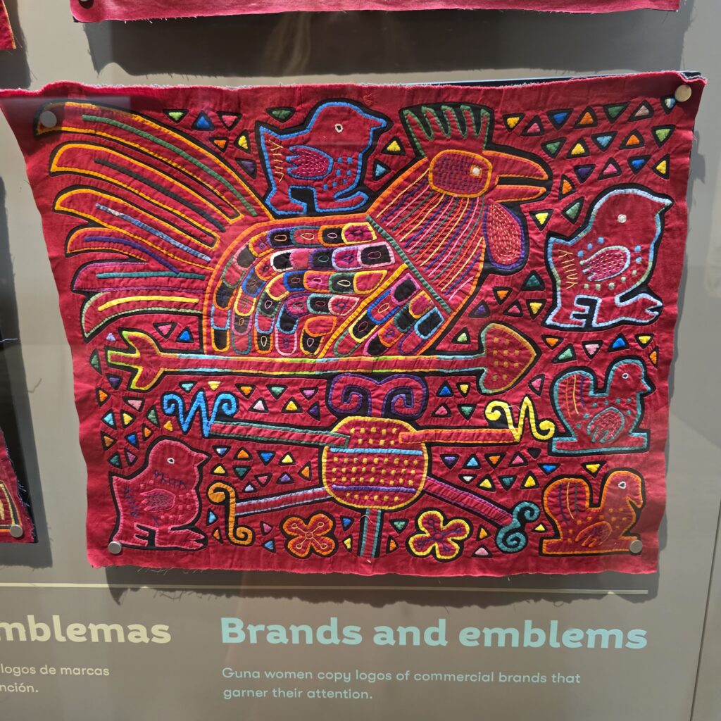 A mola with a rooster and chick depicted with a redish pink back ground and colros of blue, yellow, and green, located in the Mola Museum in Casco Viejo Panama