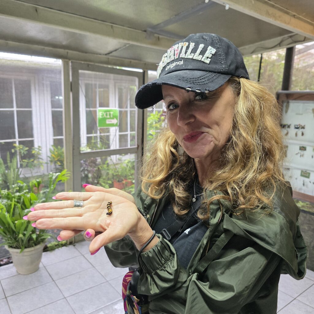 Me in a black hat and green rain jacket holding a butterfly chrysalis in the Butterfly Haven in El Valle de Anton in Panama